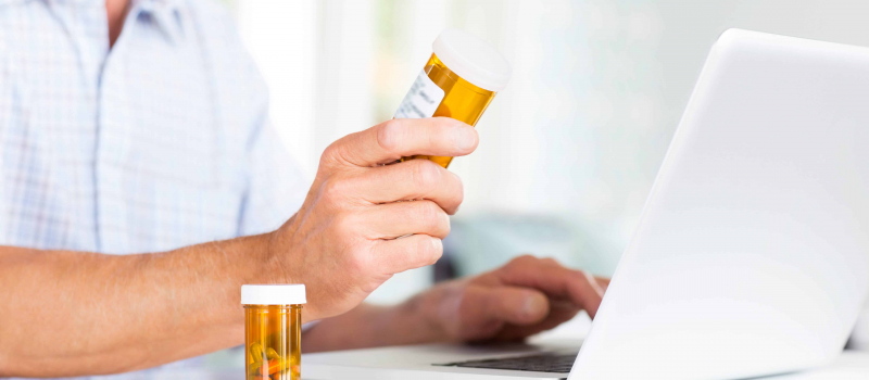 Senior Man With Pill Bottles Using Laptop At Table.